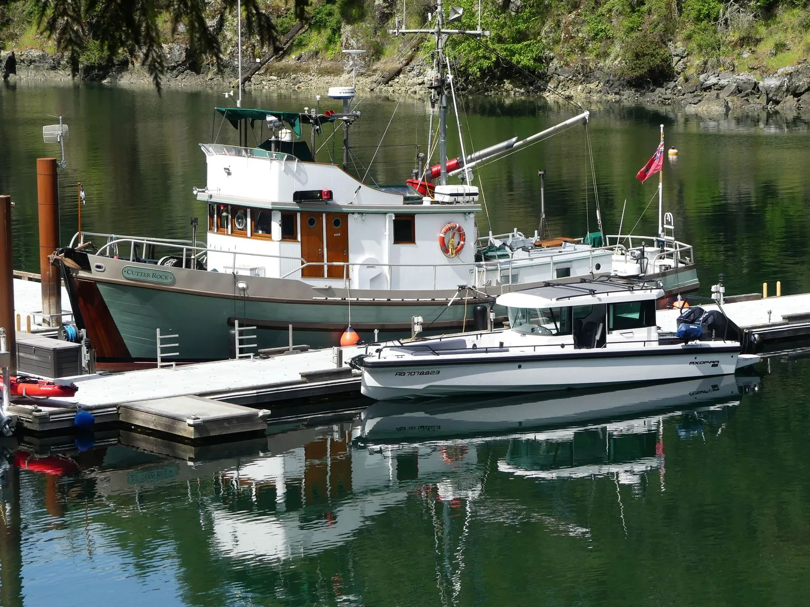Two boats docked at a peaceful harbor with picturesque reflections in the water, surrounded by nature.